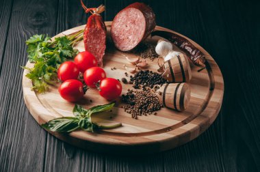 Siyah bir tablo ve bir kahverengi tahta üzerinde füme salam. Biberiye, sarımsak ve biber ve domates kiraz, dereotu, maydanoz ile sosis