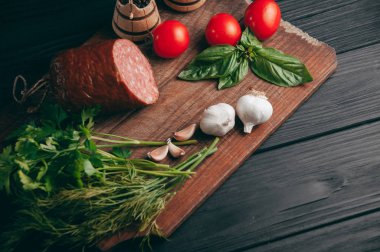 Siyah bir tablo ve bir kahverengi tahta üzerinde füme salam. Biberiye, sarımsak ve biber ve domates kiraz, dereotu, maydanoz ile sosis