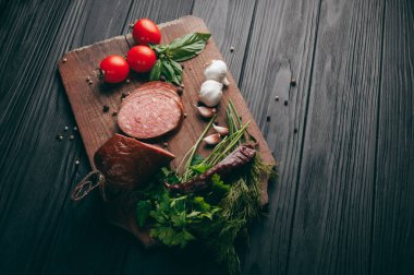 Siyah bir tablo ve bir kahverengi tahta üzerinde füme salam. Biberiye, sarımsak ve biber ve domates kiraz, dereotu, maydanoz ile sosis