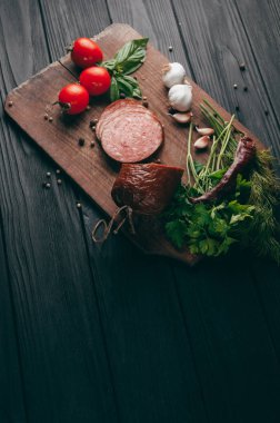 Siyah bir tablo ve bir kahverengi tahta üzerinde füme salam. Biberiye, sarımsak ve biber ve domates kiraz, dereotu, maydanoz ile sosis