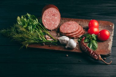 Siyah bir tablo ve bir kahverengi tahta üzerinde füme salam. Biberiye, sarımsak ve biber ve domates kiraz, dereotu, maydanoz ile sosis