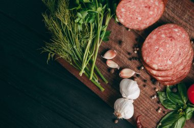 Siyah bir tablo ve bir kahverengi tahta üzerinde füme salam. Biberiye, sarımsak ve biber ve domates kiraz, dereotu, maydanoz ile sosis