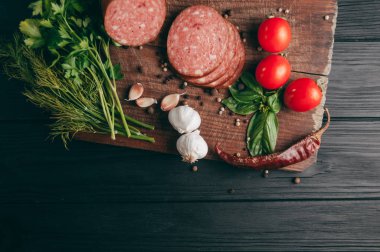 Siyah bir tablo ve bir kahverengi tahta üzerinde füme salam. Biberiye, sarımsak ve biber ve domates kiraz, dereotu, maydanoz ile sosis