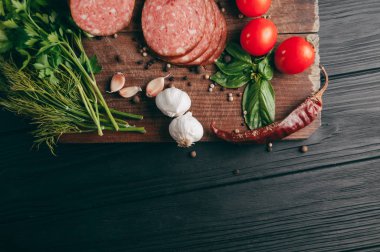 Siyah bir tablo ve bir kahverengi tahta üzerinde füme salam. Biberiye, sarımsak ve biber ve domates kiraz, dereotu, maydanoz ile sosis