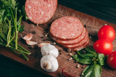 Siyah bir tablo ve bir kahverengi tahta üzerinde füme salam. Biberiye, sarımsak ve biber ve domates kiraz, dereotu, maydanoz ile sosis