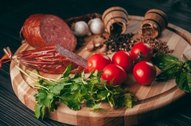 Siyah bir tablo ve bir kahverengi tahta üzerinde füme salam. Biberiye, sarımsak ve biber ve domates kiraz, dereotu, maydanoz ile sosis