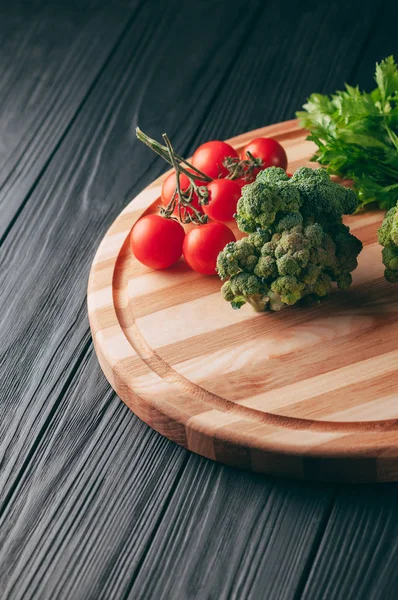On a dark wooden table, on a round wooden board fresh green broccoli, parsley, dill and cherry tomatoes for your health. Recipe. Ingredients. Dietary food. Place under the text. View from above