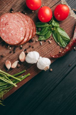 Siyah bir tablo ve bir kahverengi tahta üzerinde füme salam. Biberiye, sarımsak ve biber ve domates kiraz, dereotu, maydanoz ile sosis