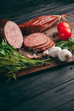 Siyah bir tablo ve bir kahverengi tahta üzerinde füme salam. Biberiye, sarımsak ve biber ve domates kiraz, dereotu, maydanoz ile sosis