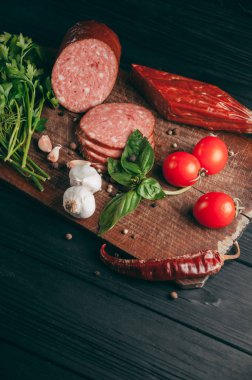 Siyah bir tablo ve bir kahverengi tahta üzerinde füme salam. Biberiye, sarımsak ve biber ve domates kiraz, dereotu, maydanoz ile sosis