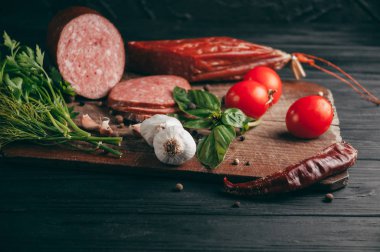 Siyah bir tablo ve bir kahverengi tahta üzerinde füme salam. Biberiye, sarımsak ve biber ve domates kiraz, dereotu, maydanoz ile sosis