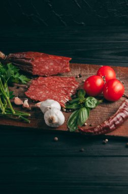 Siyah bir tablo ve bir kahverengi tahta üzerinde füme salam. Biberiye, sarımsak ve biber ve domates kiraz, dereotu, maydanoz ile sosis