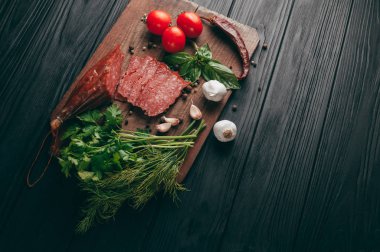 Siyah bir tablo ve bir kahverengi tahta üzerinde füme salam. Biberiye, sarımsak ve biber ve domates kiraz, dereotu, maydanoz ile sosis