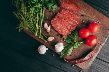 Siyah bir tablo ve bir kahverengi tahta üzerinde füme salam. Biberiye, sarımsak ve biber ve domates kiraz, dereotu, maydanoz ile sosis