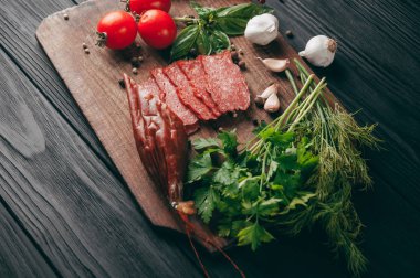 Siyah bir tablo ve bir kahverengi tahta üzerinde füme salam. Biberiye, sarımsak ve biber ve domates kiraz, dereotu, maydanoz ile sosis