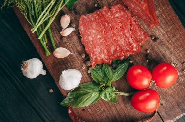 Siyah bir tablo ve bir kahverengi tahta üzerinde füme salam. Biberiye, sarımsak ve biber ve domates kiraz, dereotu, maydanoz ile sosis.