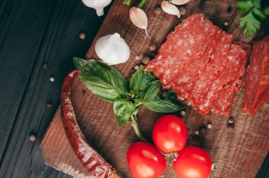 Siyah bir tablo ve bir kahverengi tahta üzerinde füme salam. Biberiye, sarımsak ve biber ve domates kiraz, dereotu, maydanoz ile sosis.