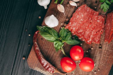 Siyah bir tablo ve bir kahverengi tahta üzerinde füme salam. Biberiye, sarımsak ve biber ve domates kiraz, dereotu, maydanoz ile sosis