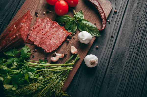 Копченая салями на черном столе и коричневой доске. Сосиски с розмарином, чесноком и перцем, томатная вишня, укроп, петрушка
