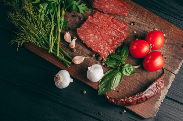 Копченая салями на черном столе и коричневой доске. Сосиски с розмарином, чесноком и перцем, томатная вишня, укроп, петрушка
