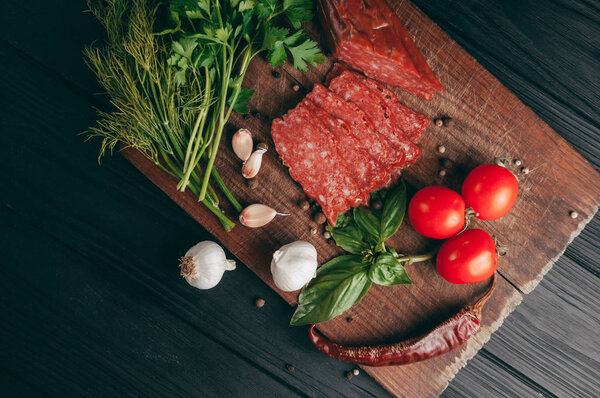 Копченая салями на черном столе и коричневой доске. Сосиски с розмарином, чесноком и перцем, томатная вишня, укроп, петрушка
