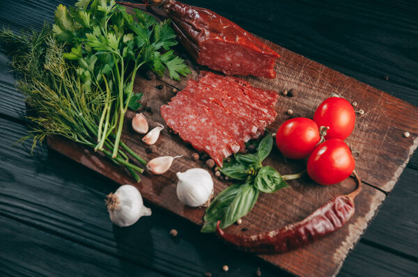 Копченая салями на черном столе и коричневой доске. Сосиски с розмарином, чесноком и перцем, томатная вишня, укроп, петрушка
.