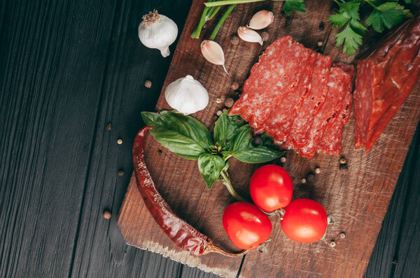 Копченая салями на черном столе и коричневой доске. Сосиски с розмарином, чесноком и перцем, томатная вишня, укроп, петрушка
.