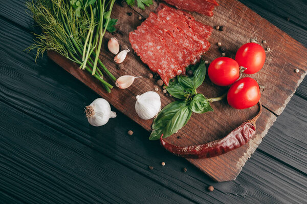 Копченая салями на черном столе и коричневой доске. Сосиски с розмарином, чесноком и перцем, томатная вишня, укроп, петрушка
.