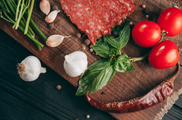 Копченая салями на черном столе и коричневой доске. Сосиски с розмарином, чесноком и перцем, томатная вишня, укроп, петрушка
.