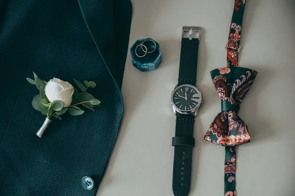 Groom's accessories: a watch, a green jacket, a buttonhole, a butterfly and rings on a tinted beige cushion