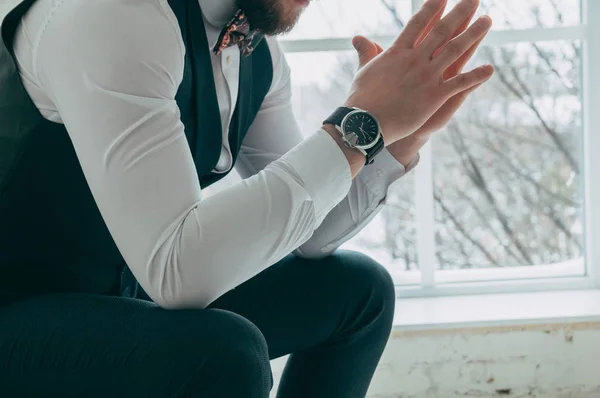 The cut-off view of the boss sitting on a chair against the background of a window holding hands together, on his hand is an expensive and stylish watch, on the background of a foot, side view