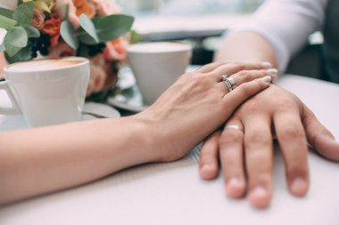 Gelin ve damat, düğün buket arka plan ve iki bardak kahve beyaz bir tablo üzerinde pahalı Alyans beyaz altın, tutan eller