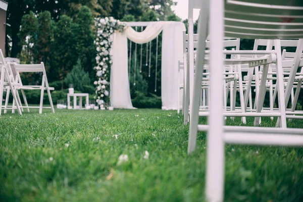 Beautiful wedding set up. Area of the wedding ceremony. Round arch, white chairs decorated with flowers, greenery. Cute, trendy rustic decor. Part of the festive decor, floral arrangement.