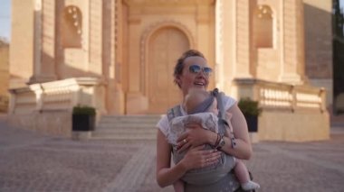 Mutlu genç bir anne Malta'da güneşli meydanında Rapide içinde bir askı içinde küçük bir bebek dans