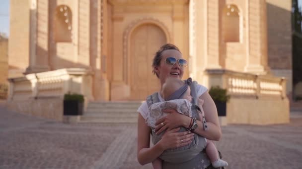 Een gelukkige jonge moeder dansen in rapide met een kleine baby in een draagdoek op het zonnige plein in Malta — Stockvideo