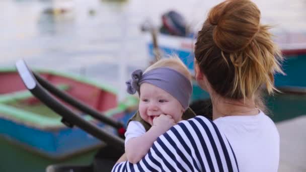 Un niño recién nacido royendo dedos, una madre sentada en el banco y mirando hacia otro lado con su hija de un año cerca del mar — Vídeo de stock