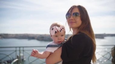 Siyah tişörtlü genç bir kadın, küçük bir bebekle birlikte denize yakın yürüyor.