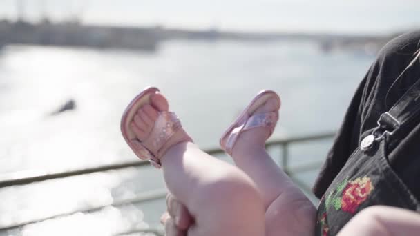 Nahaufnahme eines Kinderbeins in rosa Sandalen, eine Mutter hält ein kleines Baby in der Nähe der Meeresküste auf Händen — Stockvideo