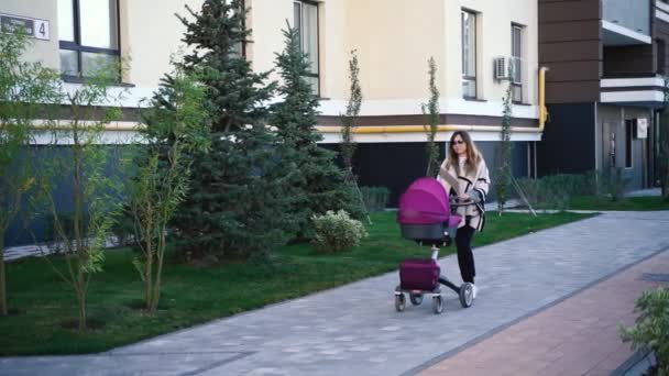 一位穿着暖和外套和白色运动鞋的年轻女子穿着慢速的婴儿车走在房子附近 — 图库视频影像