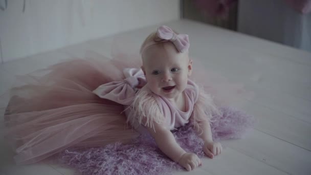 En liten baby i en rosa klänning poserar för fotot på den lila täcka och svänger de ben i slow motion — Stockvideo