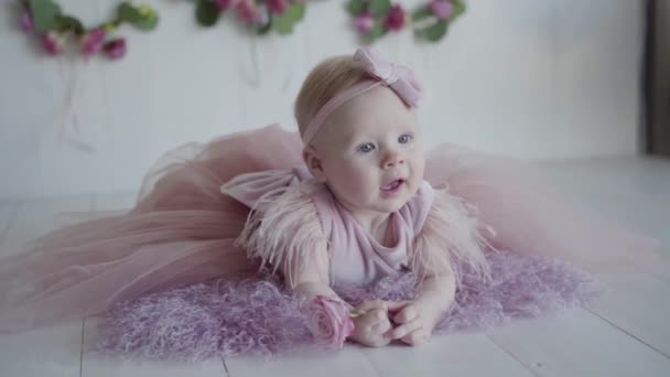 En fotosession av en liten baby i en rosa klänning liggande på magen på den lila täcka och hålla en ros — Stockvideo