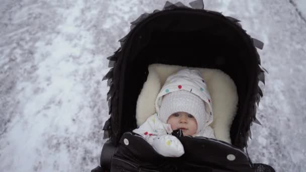 En mellersta plan om en liten flicka som sitter i en svart barnvagn. En vinterpromenad med en förälder. Slow motion. — Stockvideo