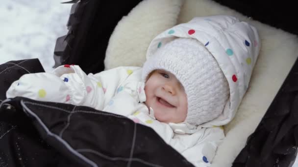 Een close-up plan van een lachende baby in de warme muts zitten in een zwarte kinderwagen in een winter park — Stockvideo