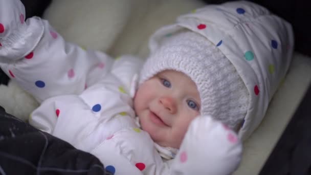 Планування крупним планом усміхненої дитини в білому костюмі, що лежить в чорному колясці в зимовому парку — стокове відео