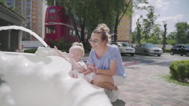 Een baby met een haar staart glimlacht in de buurt van de fontein met een jonge moeder — Stockvideo