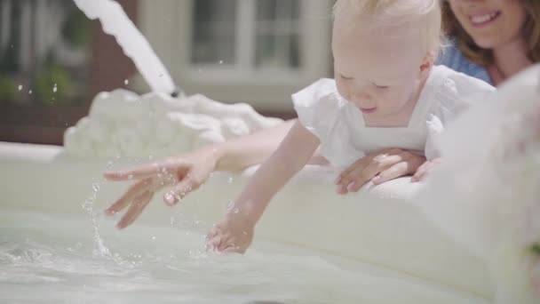 En liten flicka med en hår svans ler nära fontänen med en mor — Stockvideo