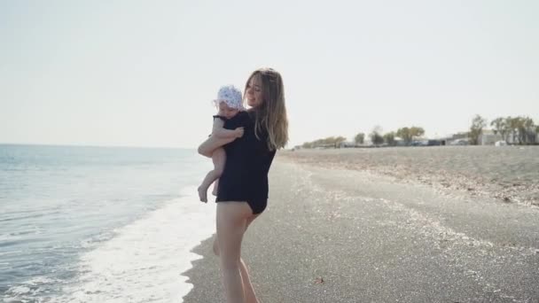 Femme souriante marche le long de la côte de la mer avec sa fille sur les mains — Video