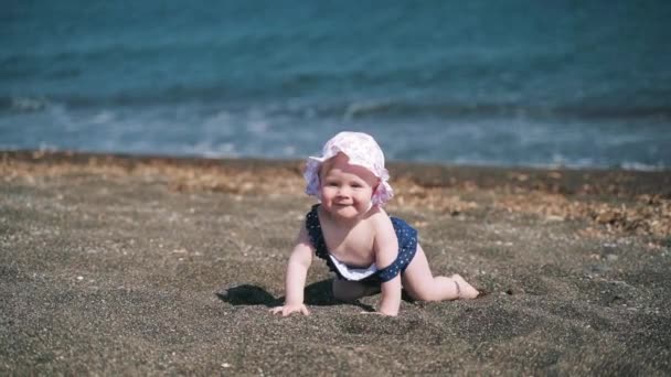 Усміхнена однорічна дівчина повзає на чорному вулканічному піщаному пляжі на острові Санторіні, Греція — стокове відео
