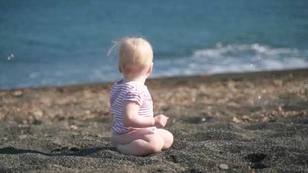 Ежегодная блондинка сидит на пляже и смотрит на пузырьки, которые летают в замедленной съемке. — стоковое видео