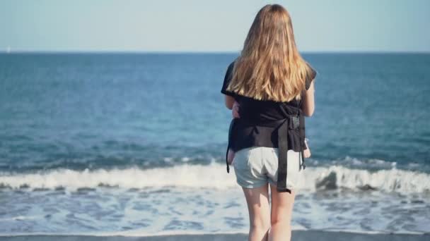 海の近くのスリングで赤ちゃんを抱いている母親の背中からのショット — ストック動画